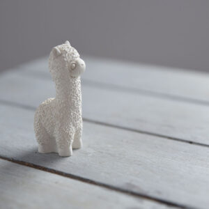 Oldalnézeti fotó egy kézműves, rusztikus textúrájú gipsz alpaka figuráról. A minimalista stílusú állatos dísztárgy egy kopottas, szürke fa felületen áll, természetes fénnyel megvilágítva.