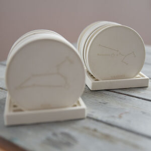 Közeli kép egy fehér gipsz poháralátétről, amelybe elegáns asztrológiai szimbólumok és csillagjegyek vannak vésve, használat közben.