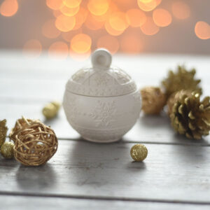 Magas szögű felvétel a zárt fehér "Karácsonyi kincsesgömb" gipsz dekorációról egy koptatott fa felületen, aranyra festett fenyőtobozokkal és fonott gömbökkel körülvéve az ünnepi hangulatért.