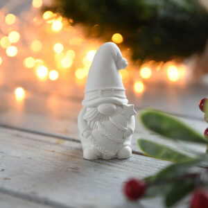 Szemből fotózott, karácsonyi dekoráció manó figura, aki egy kis fenyőfát tart. A háttérben meleg fényű égősor és fenyőágak láthatóak, kiemelve az ünnepi asztali dísz jellegét.