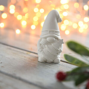 Szemből fotózott, karácsonyi dekoráció manó figura, aki egy kis fenyőfát tart. A háttérben meleg fényű égősor és fenyőágak láthatóak, kiemelve az ünnepi asztali dísz jellegét.