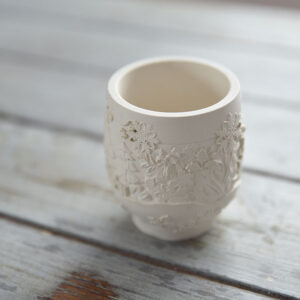 Közelkép egy kézműves beltéri gipsz dekorációról: a fénytartó felületén plasztikusan kiemelkedő téli tájkép, karácsonyfák és hókristályok láthatóak.