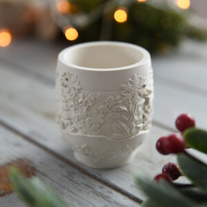 Karácsonyi dekoráció: egy henger alakú, fehér gipsz fénytartó, melynek oldalát dombornyomott hópihék és fenyőfák díszítik. Az előtérben ünnepi piros bogyós ág, a háttérben meleg fényű karácsonyi égősor látható egy rusztikus fa asztalon.