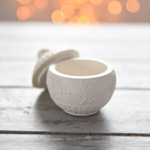 Felülnézeti kép egy nyitott, matt fehér gipsz karácsonyi kincsesgömbről, mellette a hópihe mintás tetővel. A rusztikus fehér fa asztalon aranyozott tobozok és csillogó dekorációs gömbök veszik körül.