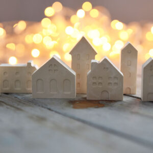 Karácsonyi dekoráció gipszből: különböző méretű fehér házikók egy rusztikus asztalon, a háttérben meleg sárga karácsonyi égősor bokeh hatású fényeivel.