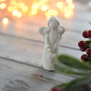 Kézzel készült fehér gipsz angyalka figura, aki egy szívet tart a kezében. A háttérben meleg fényű ünnepi világítás és piros bogyós dekoráció látható rusztikus fa felületen.