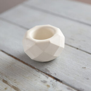 Közelkép egy geometrikus formájú, sokszögletű matt fehér gipsz mécsestartóról. A LumenStone névre keresztelt kiegészítő tiszta élekkel és sima textúrával rendelkezik, egy világosszürke rusztikus fafelületen bemutatva, tökéletes beltéri dekoráció minimál stílushoz.