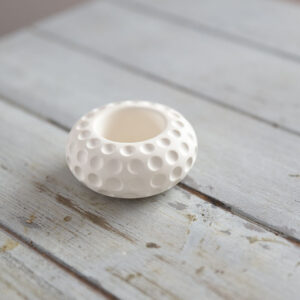 Közelkép egy kerek, matt fehér beltéri gipsz dekorációról. A LunaStone mécsestartó különlegessége a holdfelszínt idéző, mélyített pontozott textúra. A termék egy világos, rusztikus fapadlón látható, tiszta és modern stílust képviselve.