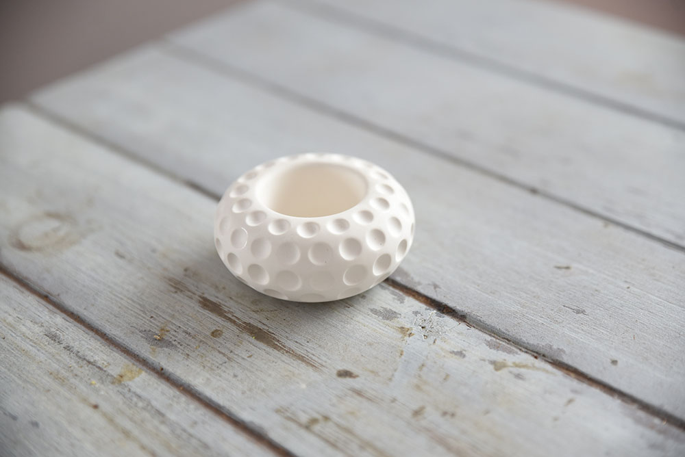 lunastone-texturalt-gipsz-mecsestarto-belteri-dekoracio Közelkép egy kerek, matt fehér beltéri gipsz dekorációról. A LunaStone mécsestartó különlegessége a holdfelszínt idéző, mélyített pontozott textúra. A termék egy világos, rusztikus fapadlón látható, tiszta és modern stílust képviselve.