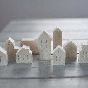 Karácsonyi dekoráció gipszből: különböző méretű fehér házikók egy rusztikus asztalon, a háttérben meleg sárga karácsonyi égősor bokeh hatású fényeivel.